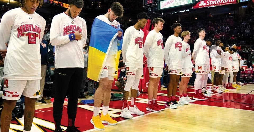 UMD men's basketball team stands on sideline, with one player with Ukrainian flag draped over his shoulders
