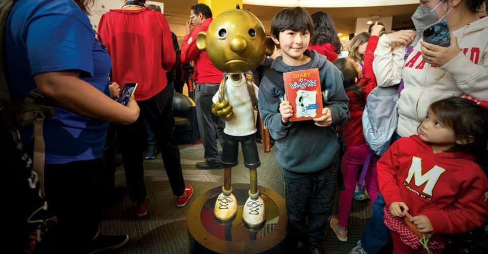Child poses with Greg Heffley statue while holding "Diary of a Wimpy Kid" book